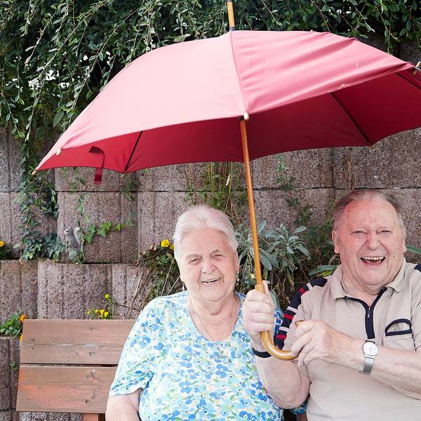 Eine alte Frau und ein alter Mann sitzen fröhlich auf einer Bank, während er einen Regenschirm hält.