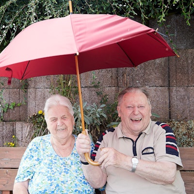 Eine alte Frau und ein alter Mann sitzen fröhlich auf einer Bank, während er einen Regenschirm hält.