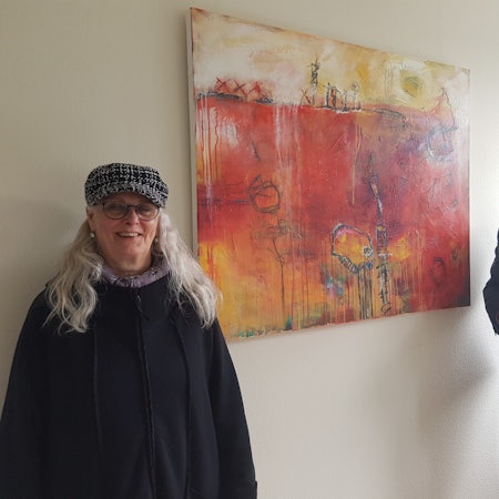 Ein Mann im blauen Sakko und Hemd neben einem abstrakten Gemälde in Rot- und Orangetönen.