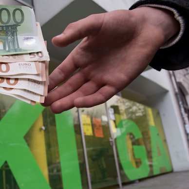 Ein Mann und eine Frau halten mehrere Geldscheine vor einem Kindergarten in den Händen.