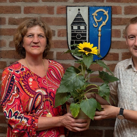 Eine Frau und ein Mann schauen in die Kamera, sie wirkt sehr ernst. Gemeinsam halten sie eine Sonnenblume in die Kamera.