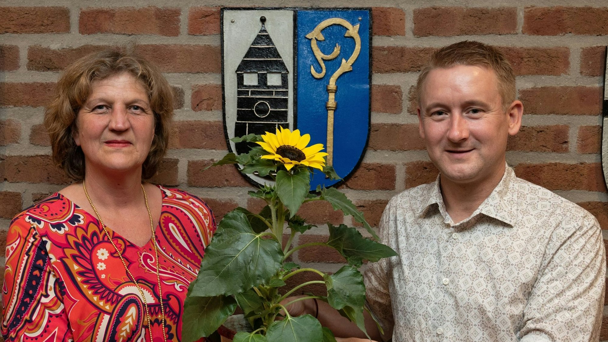 Eine Frau und ein Mann schauen in die Kamera, sie wirkt sehr ernst. Gemeinsam halten sie eine Sonnenblume in die Kamera.