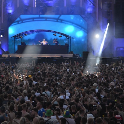 Sehr viele Menschen stehen vor einer mit hohen Elementen verkleideten Bühne, die in blaues Licht getaucht ist.