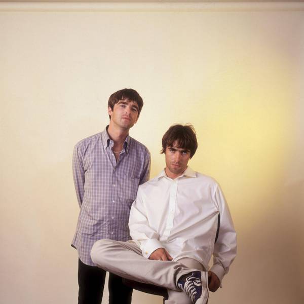 Noel Gallagher von Oasis, stehend, trägt ein kariertes Hemd, neben ihm sitzt sein jüngerer Bruder Liam im weißen Hemd.