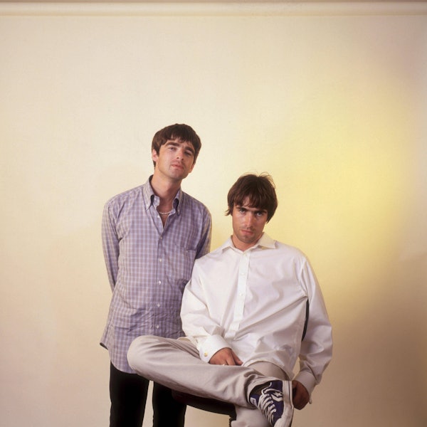 Noel Gallagher von Oasis, stehend, trägt ein kariertes Hemd, neben ihm sitzt sein jüngerer Bruder Liam im weißen Hemd.