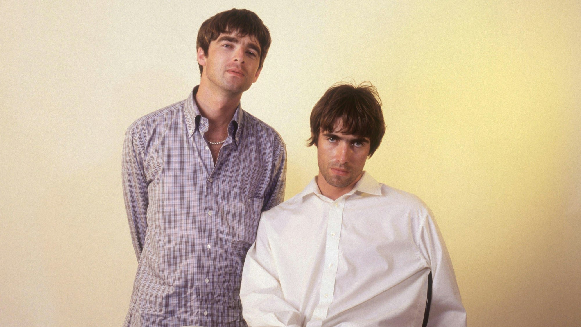 Noel Gallagher von Oasis, stehend, trägt ein kariertes Hemd, neben ihm sitzt sein jüngerer Bruder Liam im weißen Hemd.