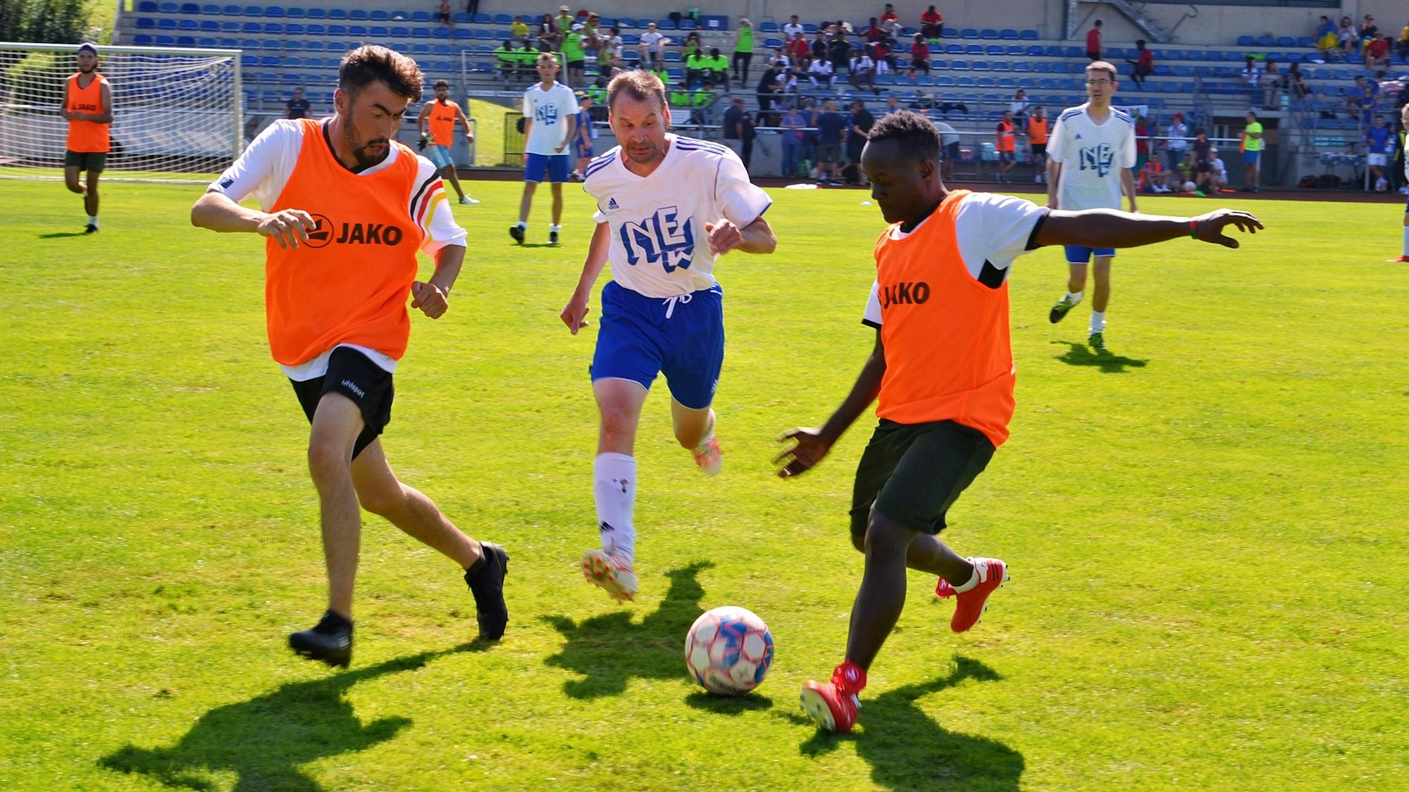 Männer spielen auf einem Rasenplatz Fußball. Im Hintergrund sind Zuschauer auf einer Tribüne zu sehen.