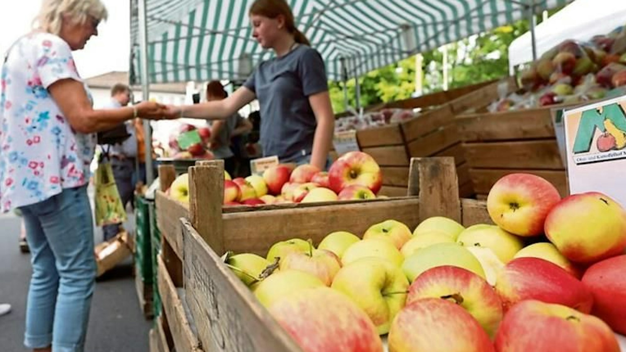 Eine Holzkiste voller Äpfel an einem Obsttand, im Hintergrund zwei Frauen ,