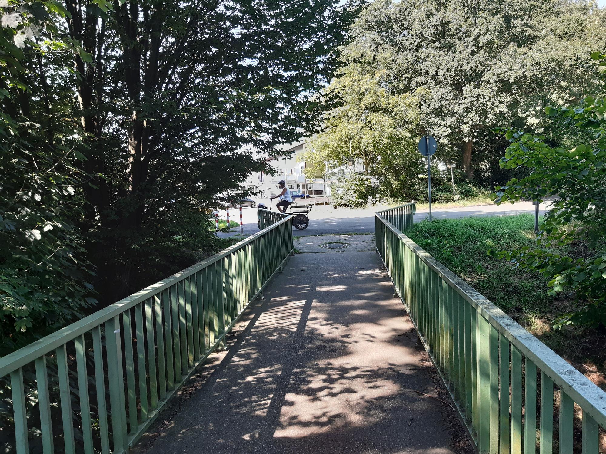 Auf dem Bild ist eine Fußgängerbrücke zu sehen, die wegen einiger Bäume links und rechts der Brücke im Halbschatten liegt.