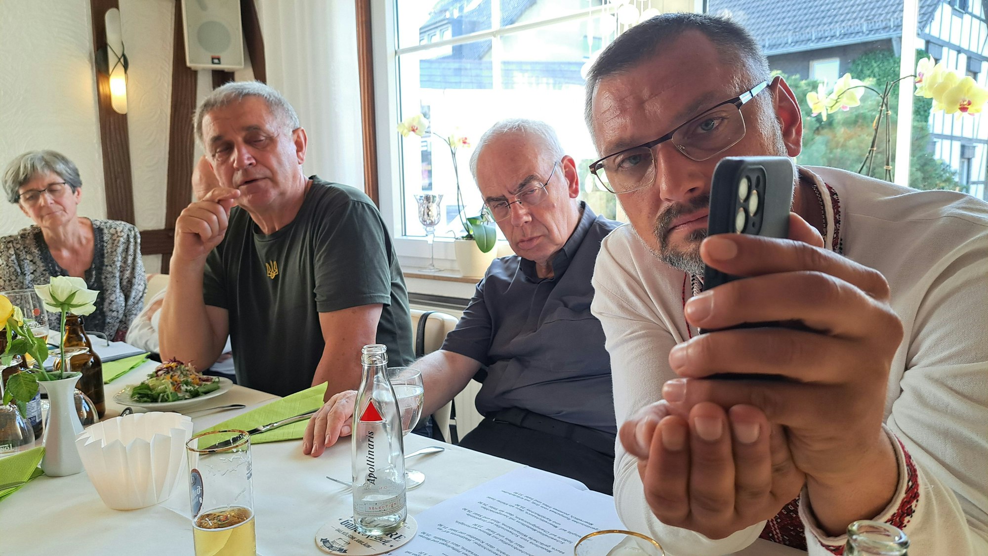 Ein Mann hält ein Smartphone, die anderen Menschen am Tisch schauen auf den Bildschirm.