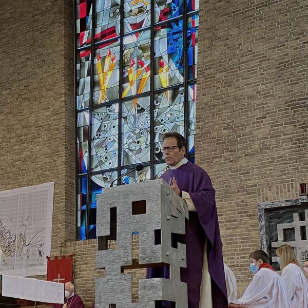 Der Pfarrer trägt eine violette Robe und steht an einem Rednerpult. Im Hintergrund ein Kirchenfenster.