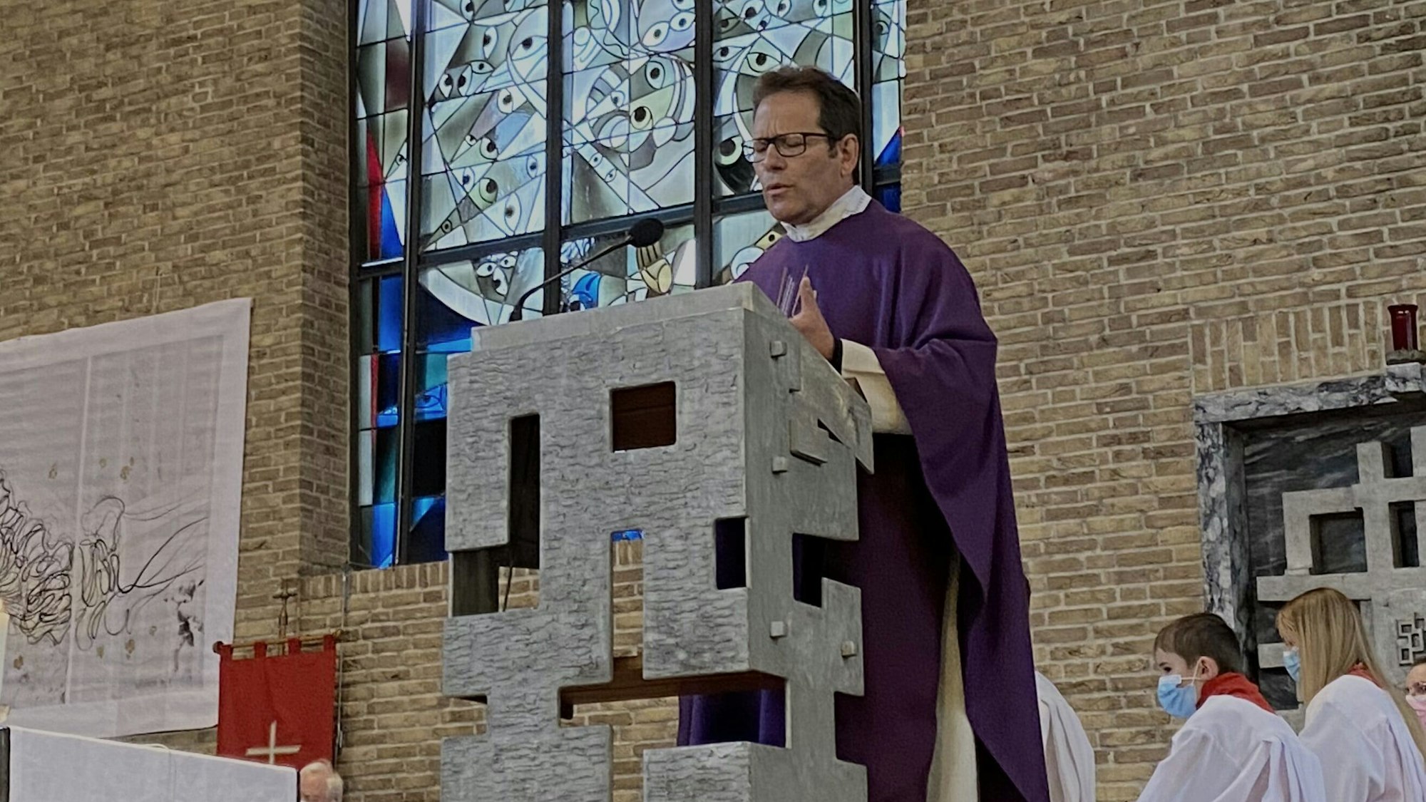 Der Pfarrer trägt eine violette Robe und steht an einem Rednerpult. Im Hintergrund ein Kirchenfenster.