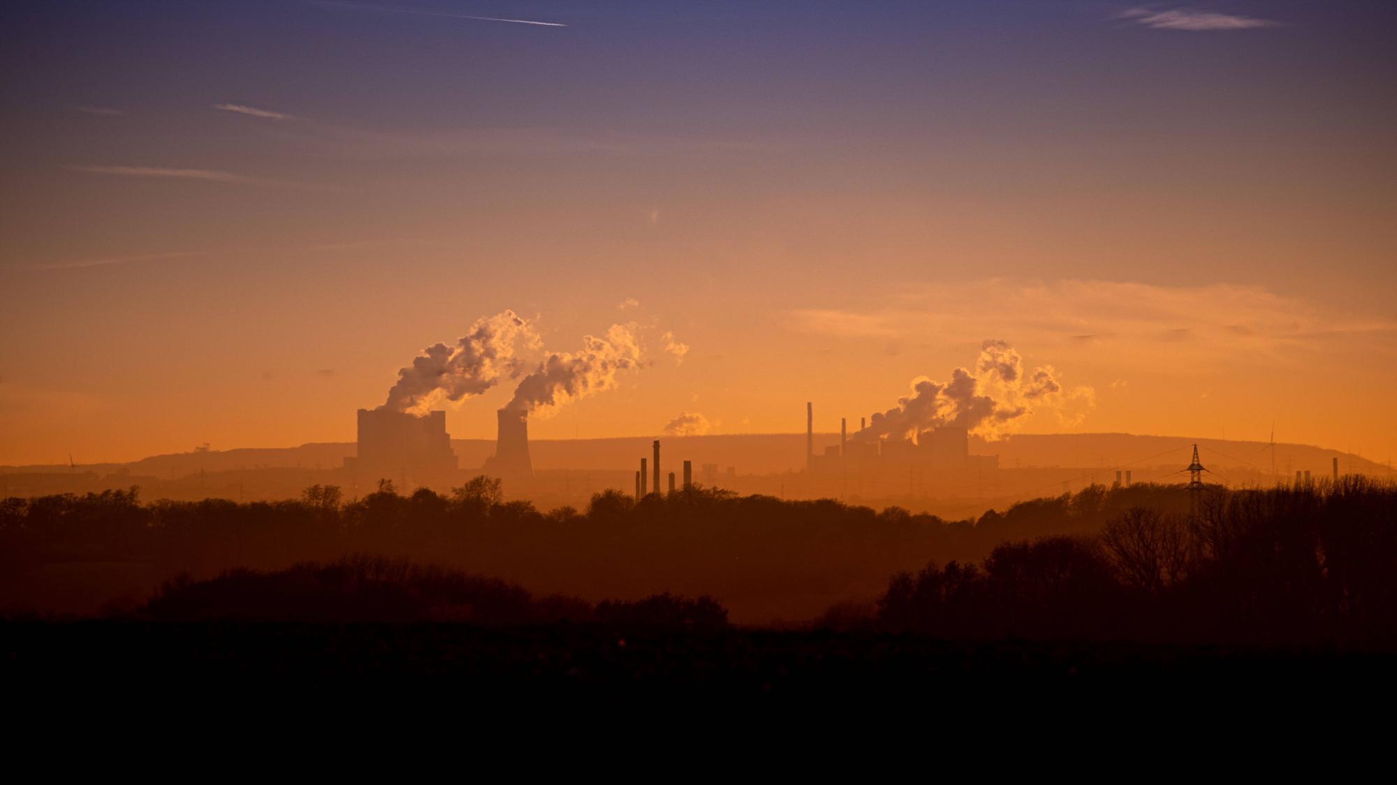 Blick auf das rheinische Braunkohlerevier in orangem Licht, mit Industrieschornsteinen und Rauch, der in den Himmel steigt.