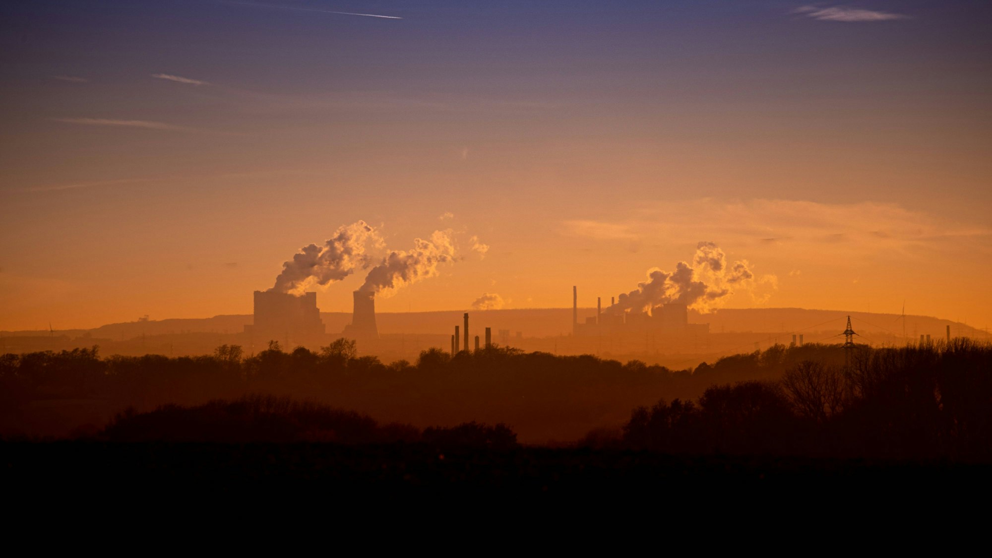 Blick auf das rheinische Braunkohlerevier in orangem Licht, mit Industrieschornsteinen und Rauch, der in den Himmel steigt.