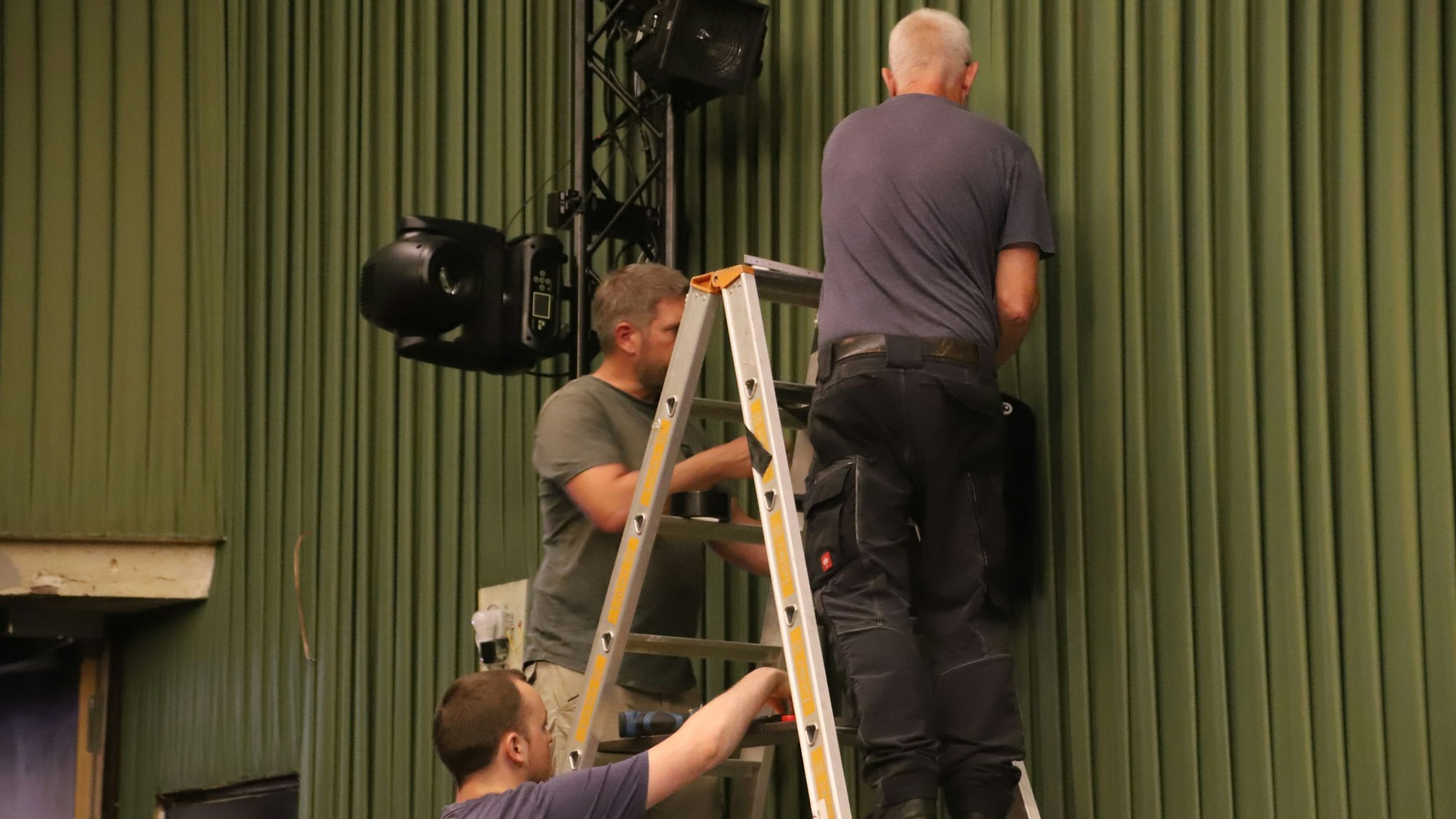 Zwei Männer auf einer Leiter hängen eine Lautsprecherbox auf, ein Dritter hilft.