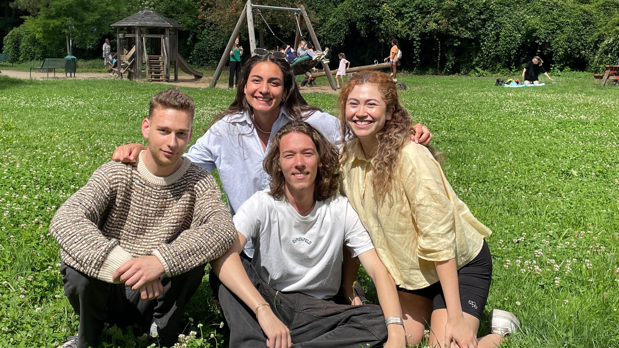 Vier junge Menschen aus Köln sitzen auf einer Wiese eines Spielplatzes, sie sind ehrenamtlich bei „youncaritas“ aktiv.