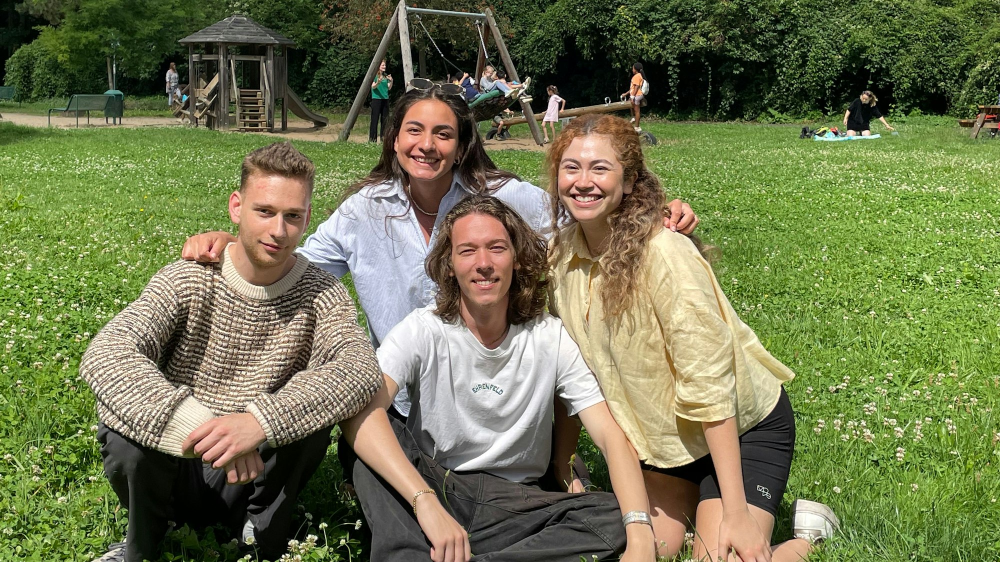 Vier junge Menschen aus Köln sitzen auf einer Wiese eines Spielplatzes, sie sind ehrenamtlich bei „youncaritas“ aktiv.