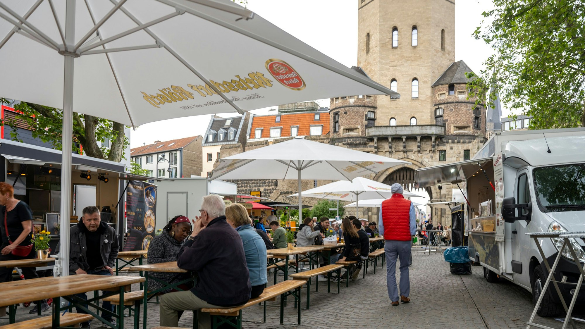 Besuchende beim Feierabendmarkt auf dem Chlodwigplatz in Köln sitzen auf Bierbänken an Tischen vor der Severinstorburg. (Archivbild)