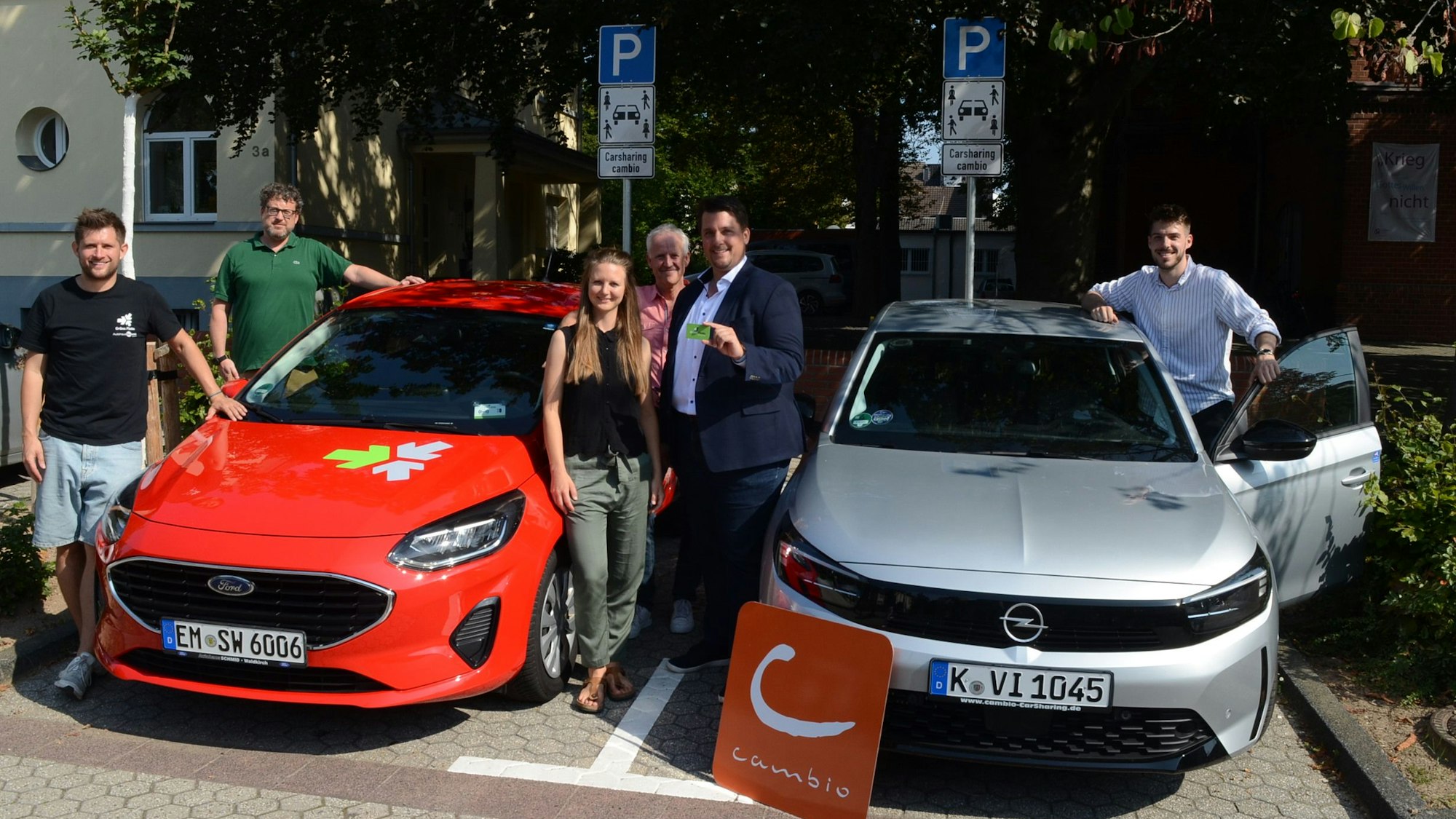 Fünf Männer und eine Frau stehen neben Kleinwagen am Rande einer Straße. Schilder weisen die Autos als Carsharing-Fahrzeuge aus.