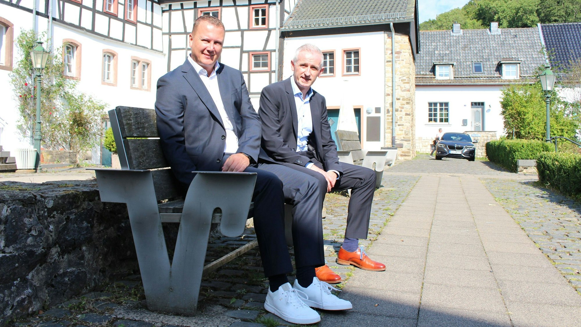 Zwei Männer in dunklen Anzügen und weißem Hemd sitzen auf einer Bank im historischen Ortskern in Blankenheim.