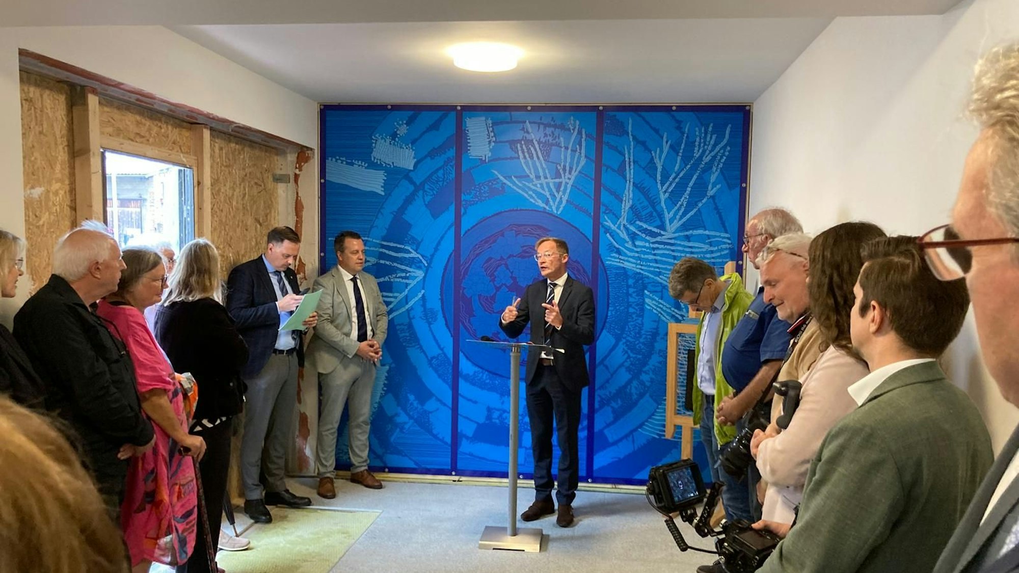 Staatssekretär Daniel Sieveke spricht vor einer in Blautönen gestalteten Wand in einem Gebäude für Kriegsgeflüchtete aus der Ukraine.