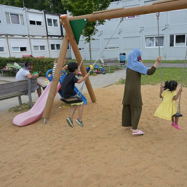 Zwei Kinder sitzen auf einer Schaukel. Eine Mutter steht dabei. Im Hintergrund sieht man die grauen Containerbauten.