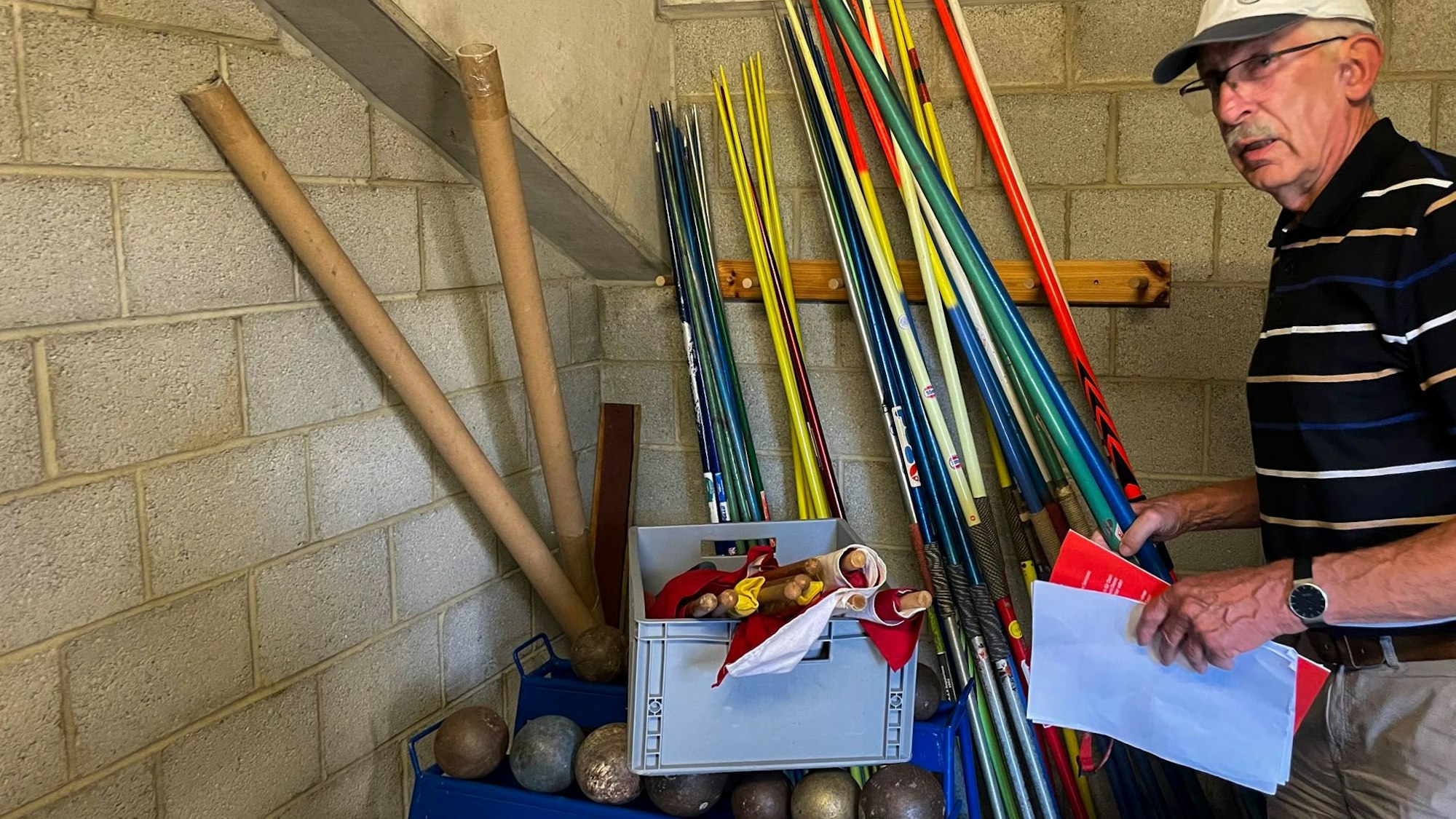 Der LGO-Vorsitzende Hans-Werner Pütz hält Zettel in der Hand und steht im Leichtathletik-Geräteraum, in dem allerhand Material liegt.