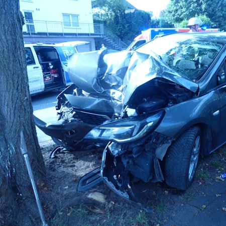 Ein Unfallauto, das nach einem Aufprall gegen einen Baum an der Frontseite völlig zerstört ist. Im Hintergrund sind Rettungskräfte zu sehen.