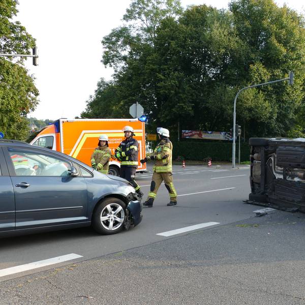 Ein schwarzes Auto liegt auf der Seite auf einer Kreuzung in Lohmar, Feuerwehrleute stehen auf der Straße.