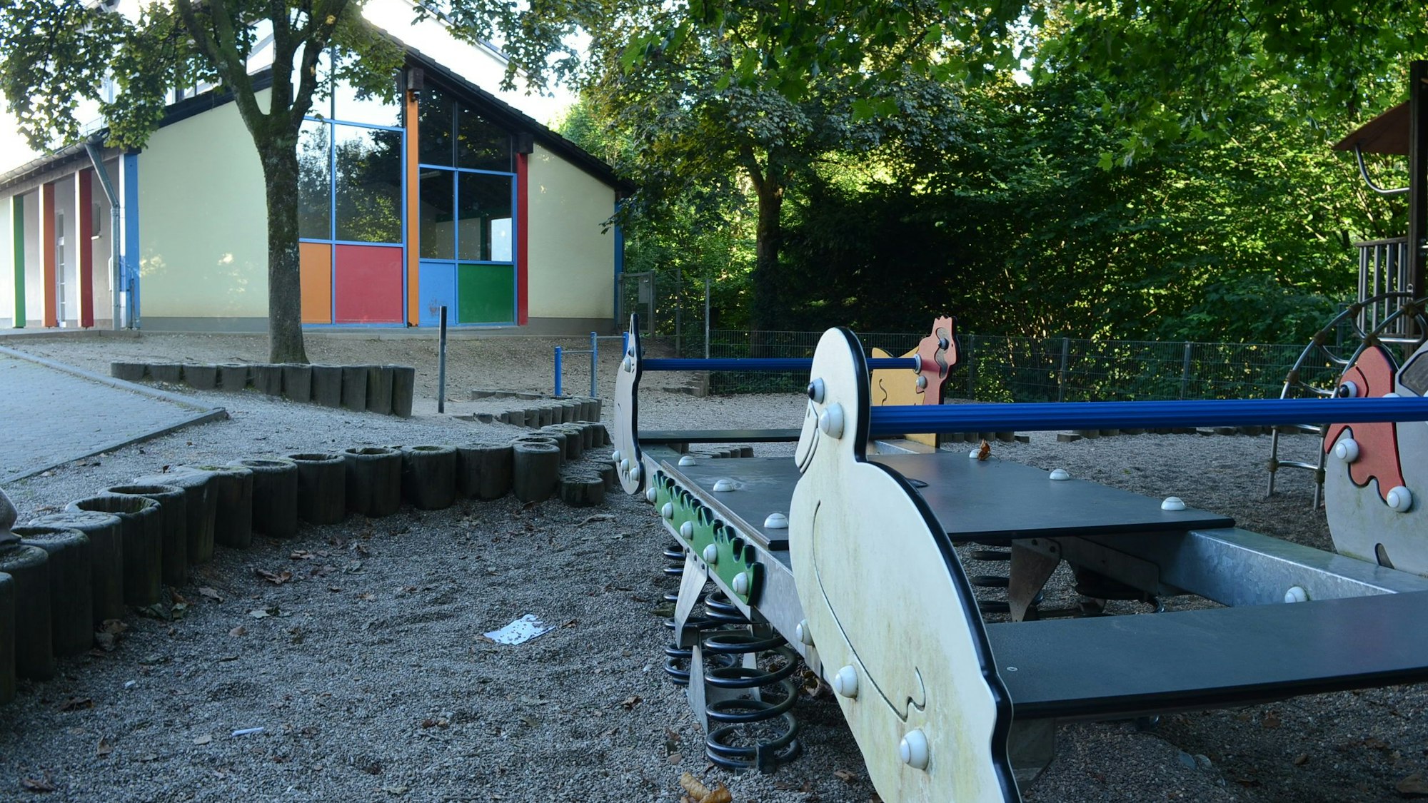 Eine Wippe und ein Klettergerüst auf einem kleinen Kinderspielplatz. Im Hintergrund steht eine seitlich offene Halle.