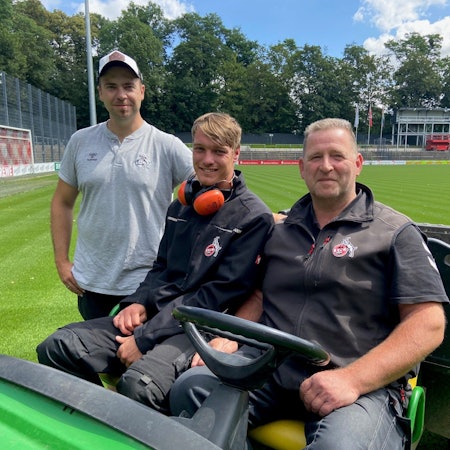 Auf dem Bild sind drei Greenkeeper auf einem Aufsitztraktor zu sehen.