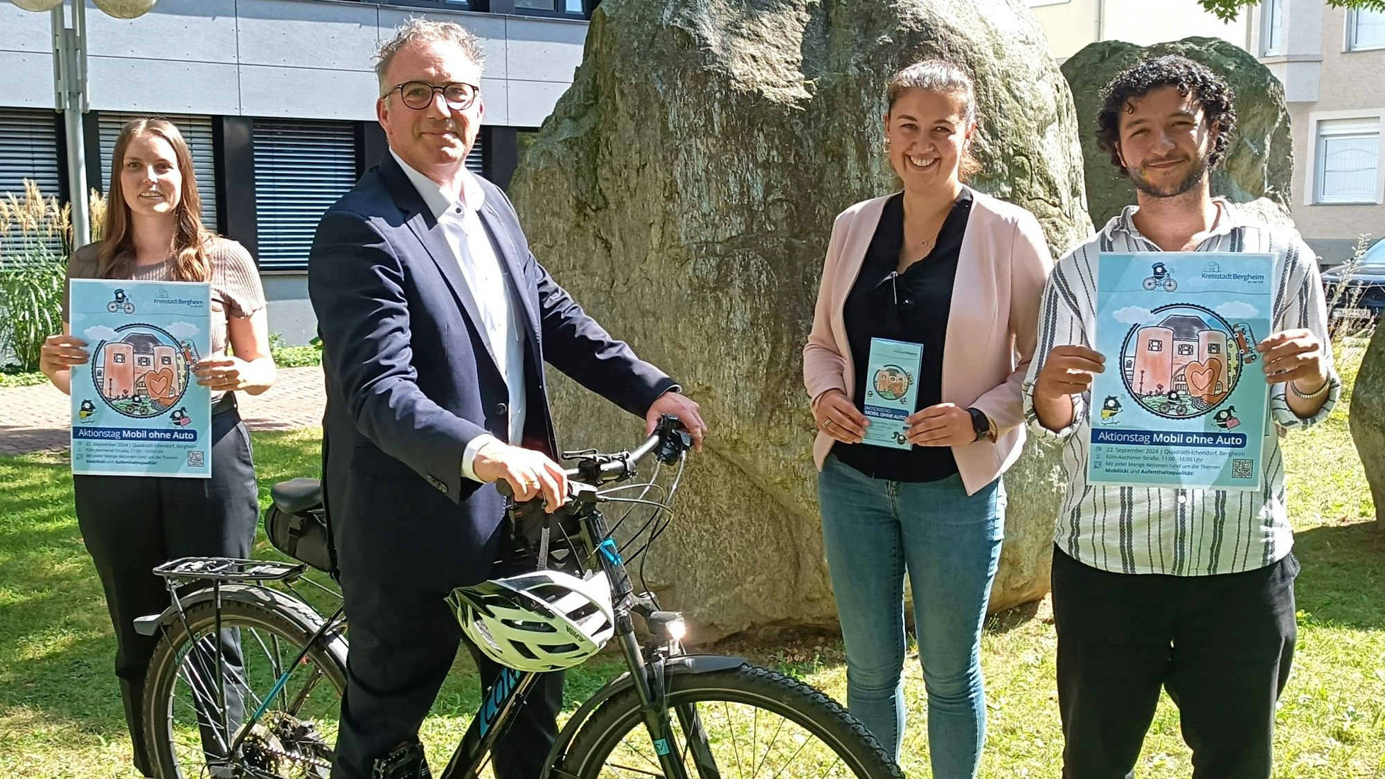 Vier Menschen stehen mit Plakaten und Flyern in den Händen und einem Fahrrad auf einer Grünfläche.