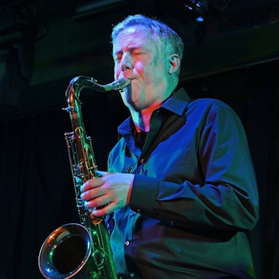 Der Musiker trägt ein dunkles Hemd und bläst in sein Saxofon, der Hintergrund ist dunkel. Vor ihm ein längliches Mikrofon.