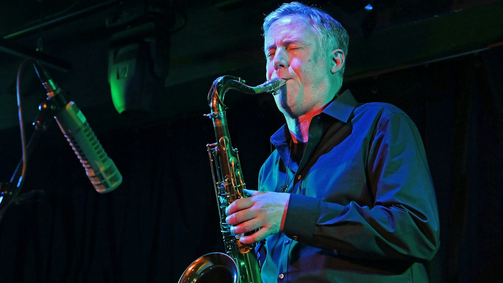 Der Musiker trägt ein dunkles Hemd und bläst in sein Saxofon, der Hintergrund ist dunkel. Vor ihm ein längliches Mikrofon.
