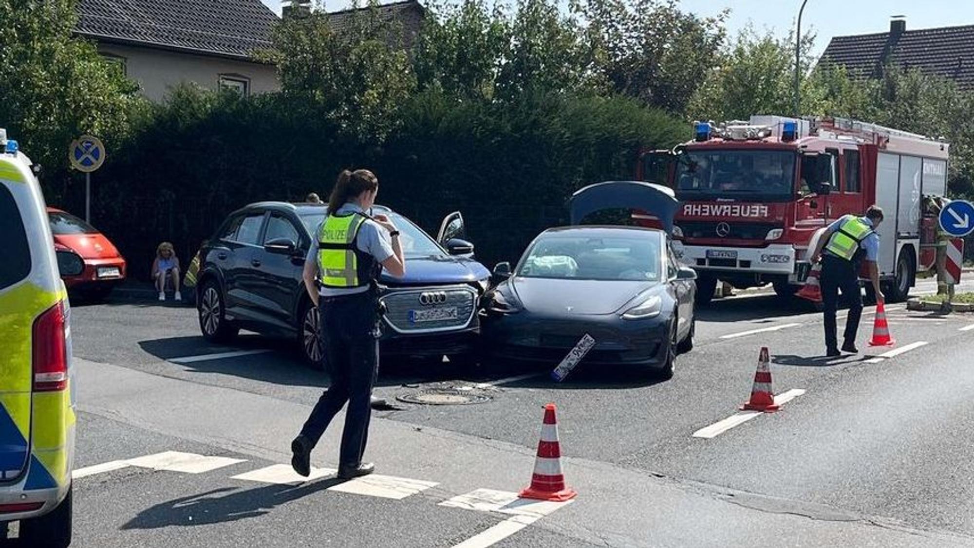 Bei einem Unfall stehen zwei kollidierte Autos nebeneinander. Daneben sind Polizeibeamte, ein Polizei- und ein Feuerwehrfahrzeug zu sehen.