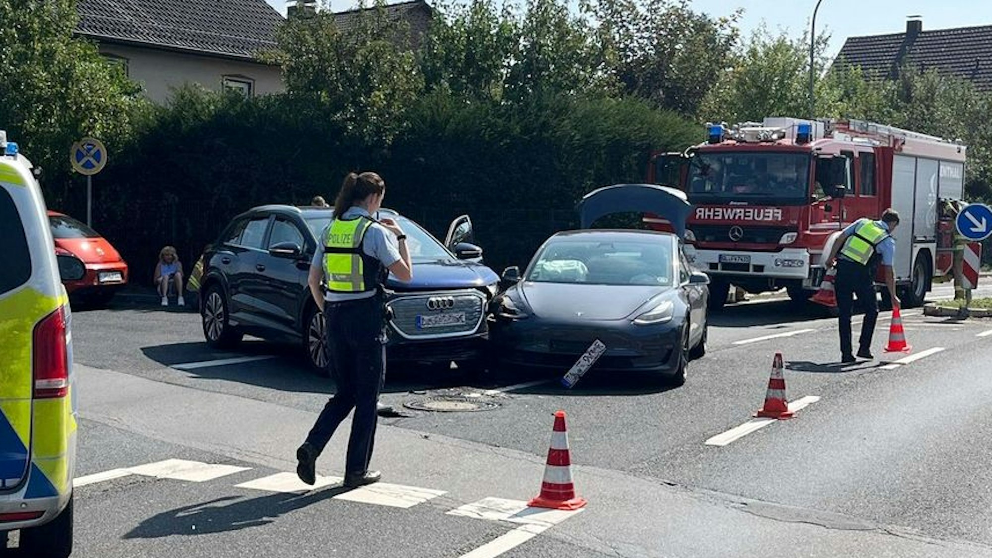 Bei einem Unfall stehen zwei kollidierte Autos nebeneinander. Daneben sind Polizeibeamte, ein Polizei- und ein Feuerwehrfahrzeug zu sehen.