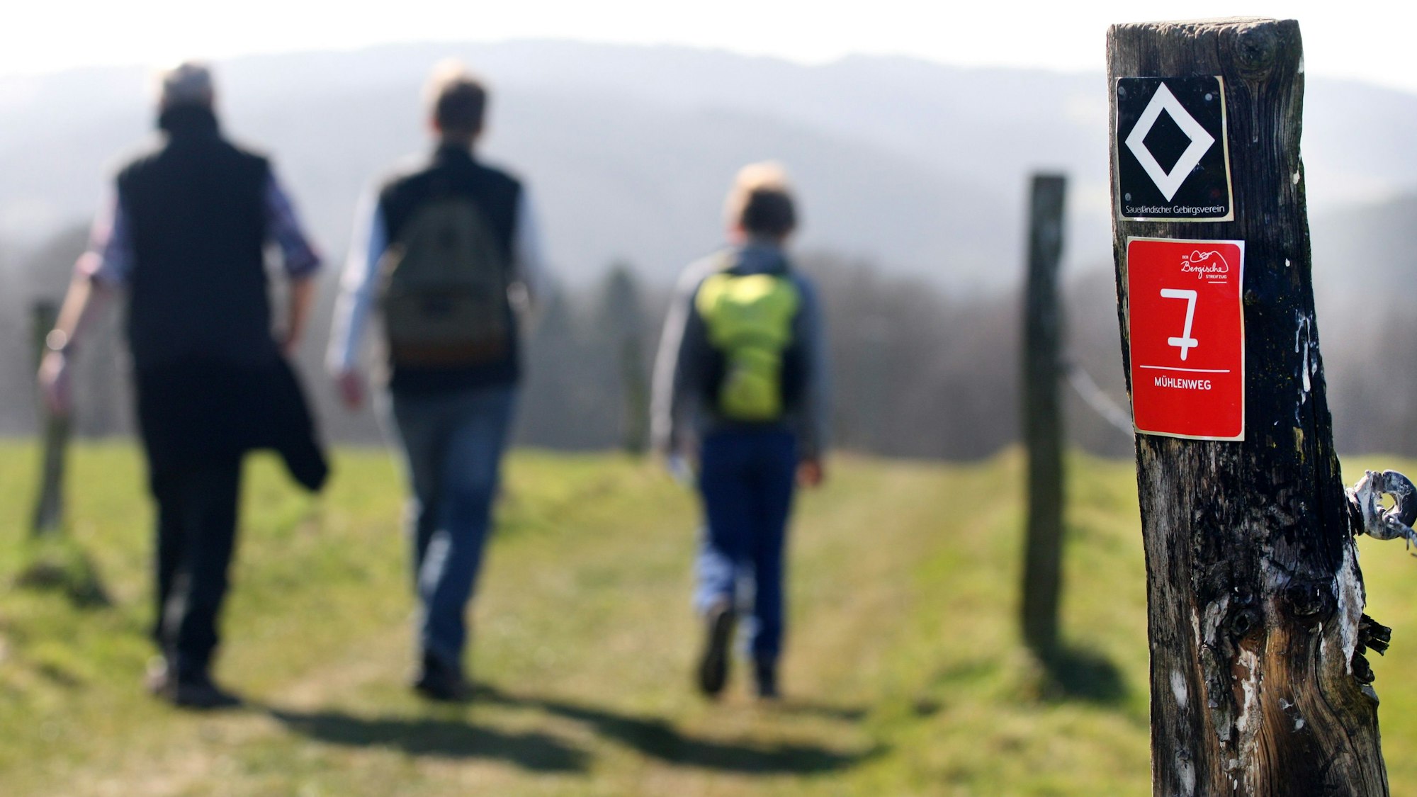 Eine Familie wandert auf dem Kürtener Mühlenweg, an einem Zaunpfosten ist eine Wegemarkierung zu sehen.