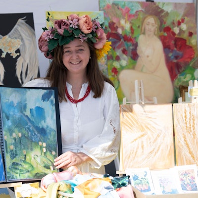 Porträt der ukrainischen Künstlerin Dara Habich: Sie zeigt zwei ihrer Bilder. Sie trägt einen Haarkranz aus bunten Blumen und lacht.