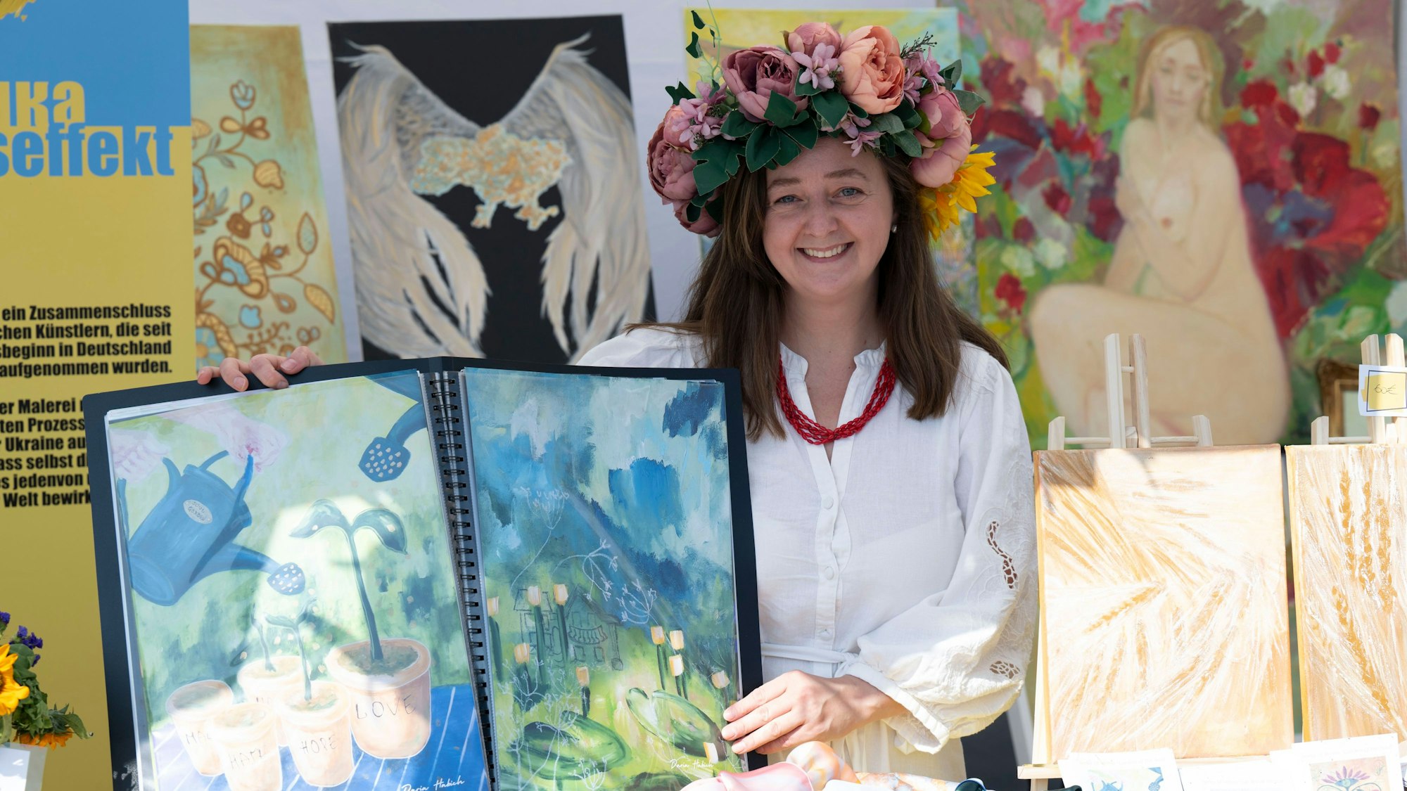 Porträt der ukrainischen Künstlerin Dara Habich: Sie zeigt zwei ihrer Bilder. Sie trägt einen Haarkranz aus bunten Blumen und lacht.