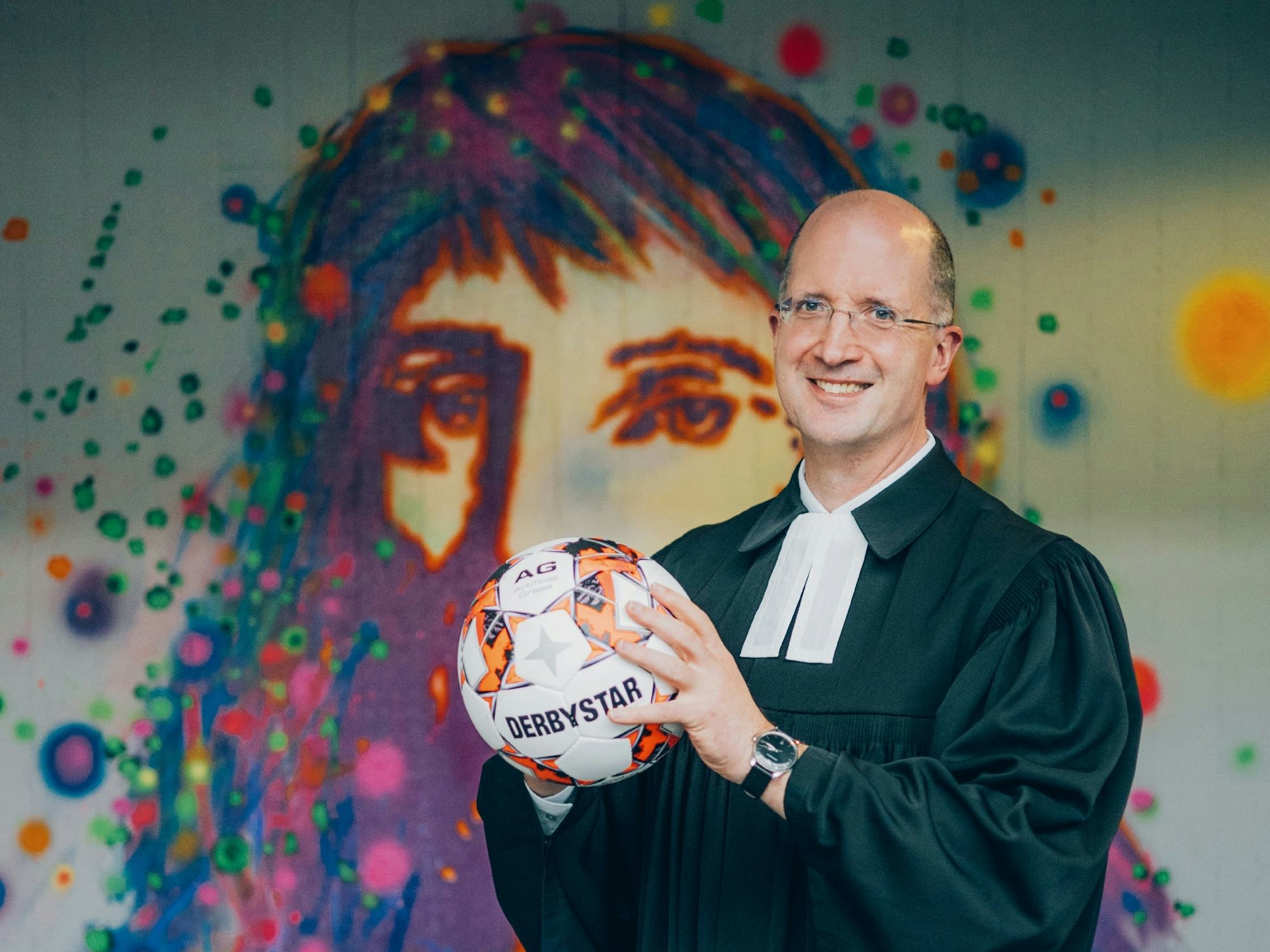 Thorsten Latzel, Präses der Evangelischen Kirche im Rheinland, steht im Talar vor einem Pop-Art-Wandbild und hält einen Fußball in den Händen. Latzel ist auch Sportbeauftragter der EKD.