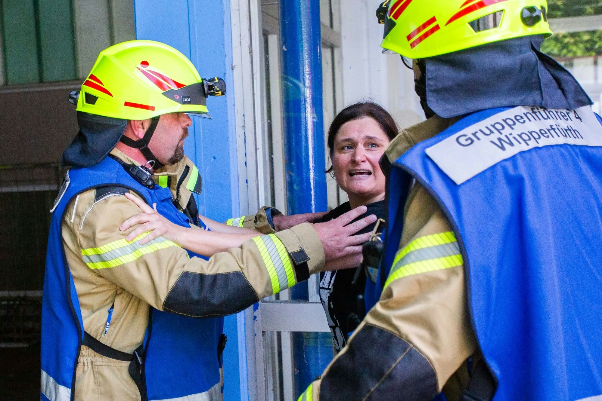 Zwei Feuerwehrleute versuchen eine aufgeregte Frau daran zu hindern, in ein Gebäude zu gelangen.