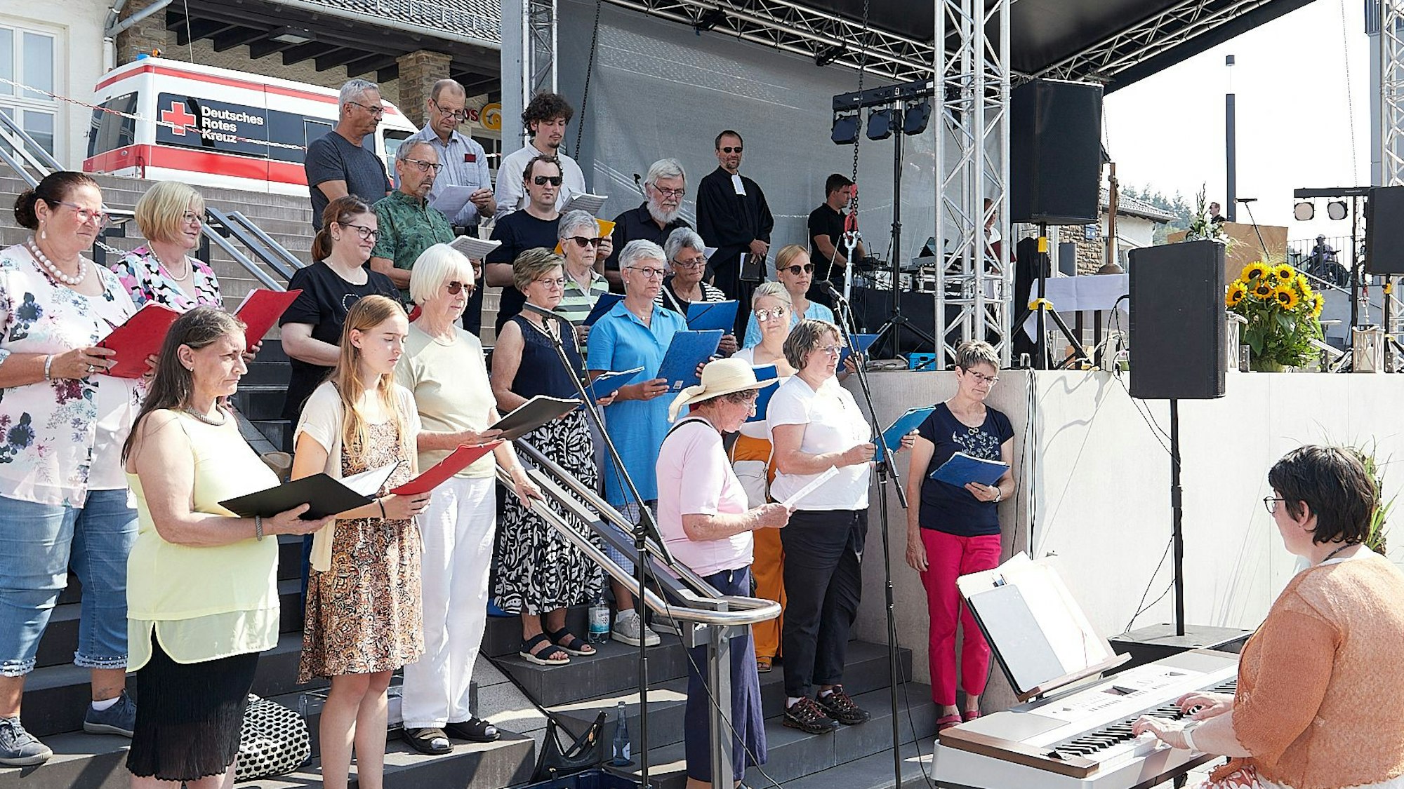 Die Mitglieder des Chores stehen auf einer Treppe und halten Notenordner in der Hand. Regionalkantorin Holle Goertz begleitet sie an einem Tasteninstrument.