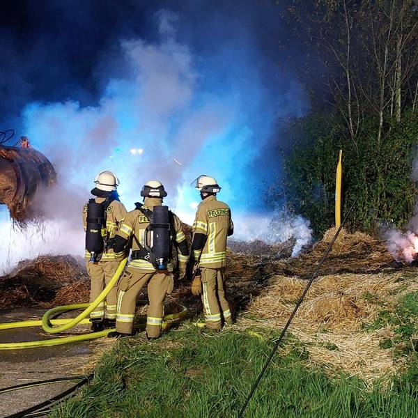 Helfer der Feuerwehr löschen Strohballen auf einer Wiese.
