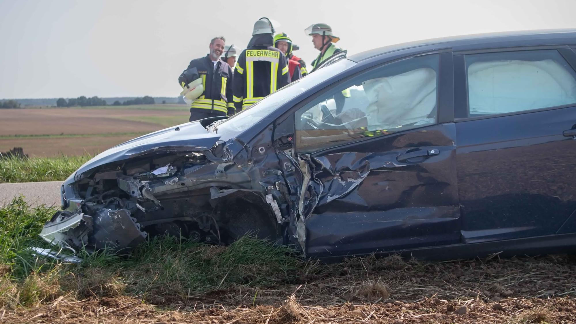 Ein Wagen, der im vorderen Bereich stark beschädigt ist, steht am Rande eines Feldes. Im Hintergrund beraten Feuerwehrleute das weitere Vorgehen.