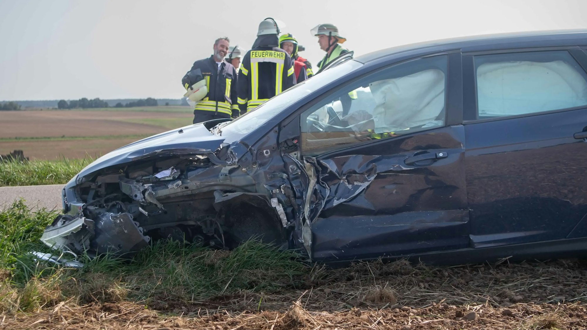 Ein Wagen, der im vorderen Bereich stark beschädigt ist, steht am Rande eines Feldes. Im Hintergrund beraten Feuerwehrleute das weitere Vorgehen.