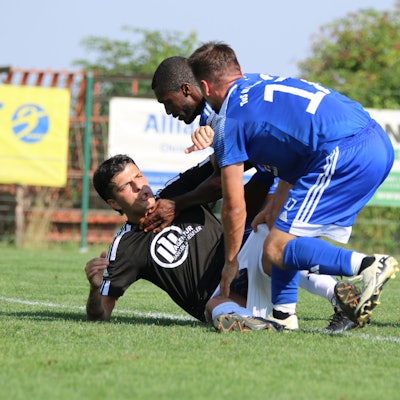Ein Fußballspieler, der auf dem Boden liegt, und zwei Gegenspieler befinden sich in einer Auseinandersetzung.
