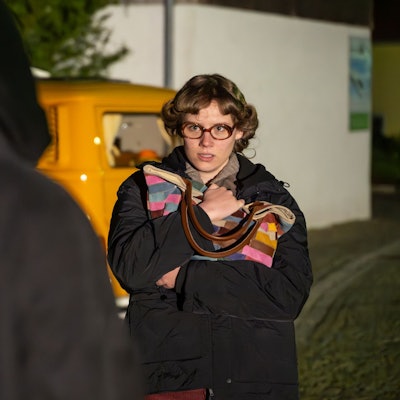 Eine junge Frau mit lockigen kurzen Haaren und Brille steht vor einem Mann in schwarzer Kleidung. Sie hält ihre Handtasche fest vor die Brust gedrückt.