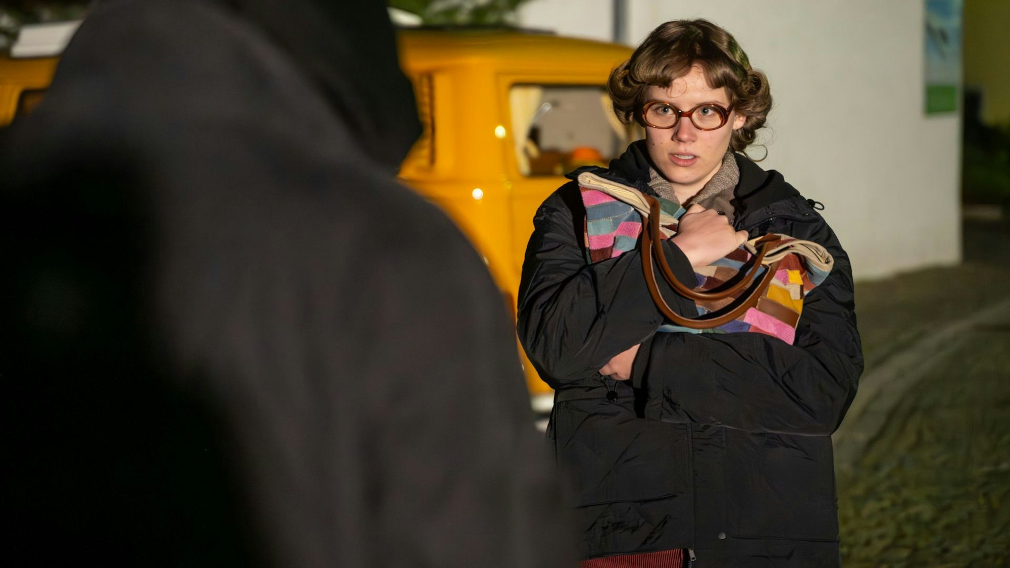 Eine junge Frau mit lockigen kurzen Haaren und Brille steht vor einem Mann in schwarzer Kleidung. Sie hält ihre Handtasche fest vor die Brust gedrückt.