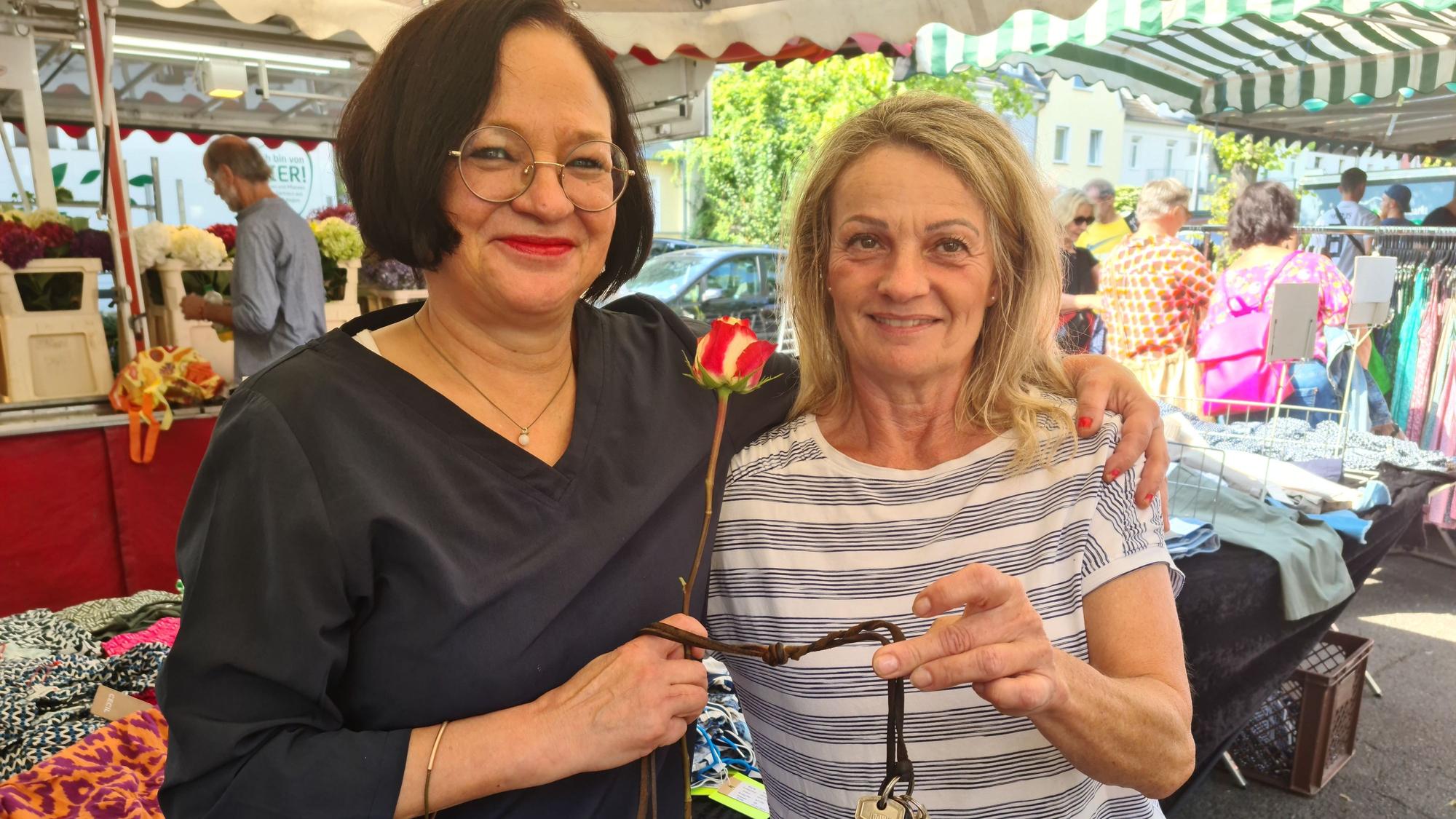 Eine dunkelhaarige Frau mit Brille überreicht einer blonden etwas kleineren Frau eine rotweiße Rose.