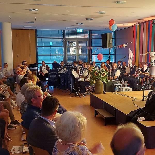 Ralf Kramp (r.) und Carsten S. Henn sitzen auf der Bühne, davor das Publikum. Die Musikkapelle Nettersheim ist hinten links platziert.